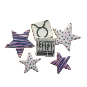 May include: Five decorated sugar cookies. Two cookies are star-shaped with pink and purple stripes. Two cookies are star-shaped with pink and purple polka dots. One cookie is square with the word "taurus" in a retro font.