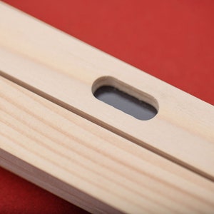 May include: Close-up of a wooden frame with a small rectangular hole cut out. The hole is centered on the frame and is surrounded by a smooth, light wood surface.