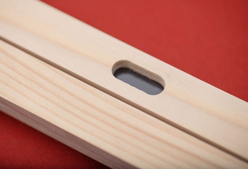 May include: Close-up of a wooden frame with a small rectangular hole cut out. The hole is centered on the frame and is surrounded by a smooth, light wood surface.