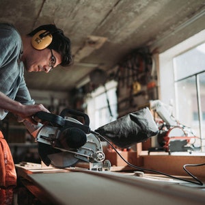May include: A person wearing ear protection uses a circular saw to cut a piece of wood in a workshop.