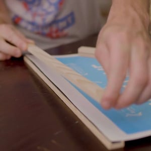 May include: A person is using tape to secure a wooden frame to a blue poster with the text "The Mill" printed on it.