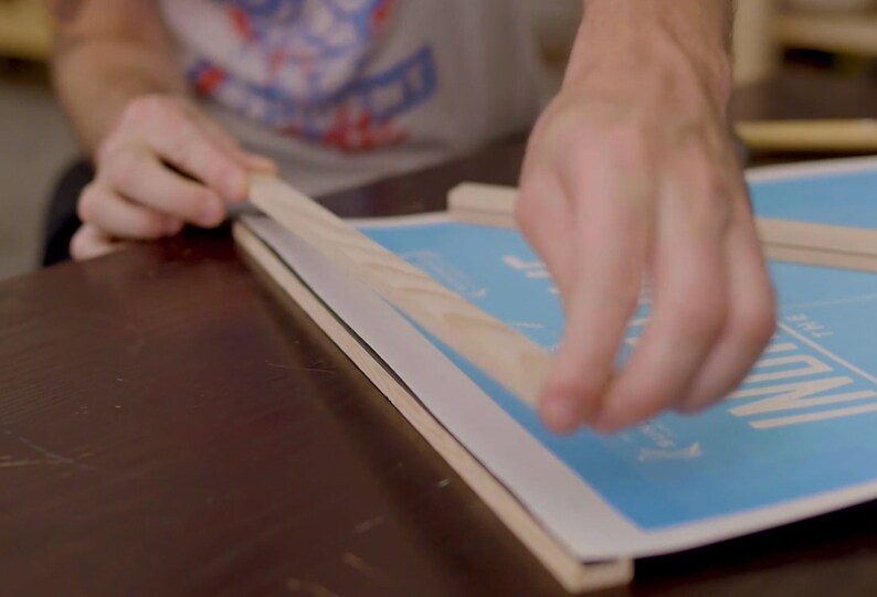 May include: A person is using tape to secure a wooden frame to a blue poster with the text "The Mill" printed on it.