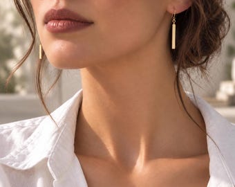 May include: A pair of gold-toned drop earrings featuring a slender, vertical rectangular bar pendant suspended from a simple hook wire. The earrings hang delicately from the earlobes, showcasing a polished metallic finish and a clean, linear design.