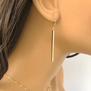 May include: Gold bar earrings with a simple design. The earrings are long and dangle from the earlobe.