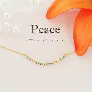 Puede incluir: Un collar de cadena dorada con cuentas turquesas y la palabra "Peace" escrita en una tarjeta blanca. El collar está sobre una superficie blanca con una flor blanca.