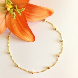 May include: A delicate gold chain necklace with small round beads. The necklace is on a white background with an orange flower.