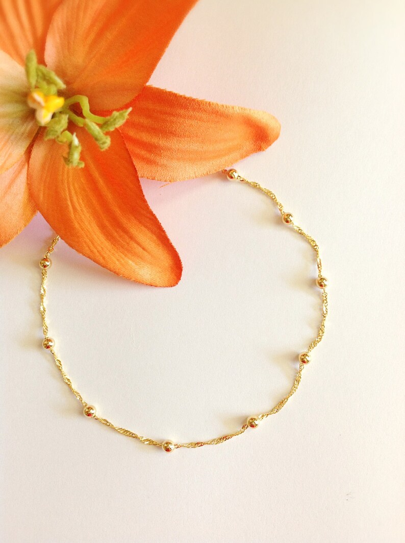 May include: A delicate gold chain necklace with small round beads. The necklace is on a white background with an orange flower.