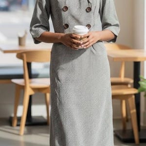 May include: A gray and white checkered dress with a collared neckline and three-quarter sleeves. The dress features a double-breasted design with brown buttons. A person is holding a white coffee cup. The background includes wooden chairs and tables.
