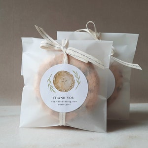 May include: Two white paper bags with a brown and white ribbon tied around them. Each bag contains a small pie and a round sticker that says "Thank you for celebrating our cutie pie!" with a watercolor illustration of a pie.
