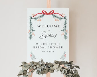 May include: A white sign with a green and red watercolor wreath, a red ribbon with a bow, and the text "Welcome to Sophie's Merry Little Bridal Shower 03.12.2024".