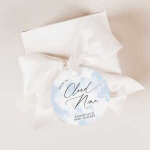 May include: A round blue and white baby shower favour tag with the text "We're on Cloud Nine" and "Samantha's Baby Shower".