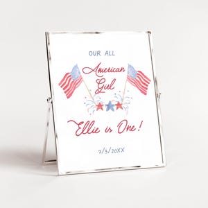 May include: A white framed sign with a red, white and blue American flag design. The text reads "Our All American Girl Ellie is One! 7/3/20XX".