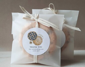 Op de afbeelding: Twee witte cadeauzakjes met een bruin lint en een rond bedankkaartje. Het kaartje heeft een aquarel-illustratie van twee mini-taarten en de tekst "THANK YOU for celebrating our cutie pie!"