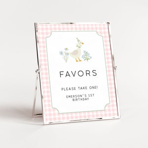 May include: A pink and white gingham favor sign with a cartoon duck wearing a party hat. The sign says "Favors, Please take one! Emerson's 1st Birthday."