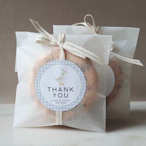 May include: White favor bags with a blue and white gingham circle sticker with a cartoon goose wearing a party hat. The sticker says "Thank You" and "Charlie's First Birthday".