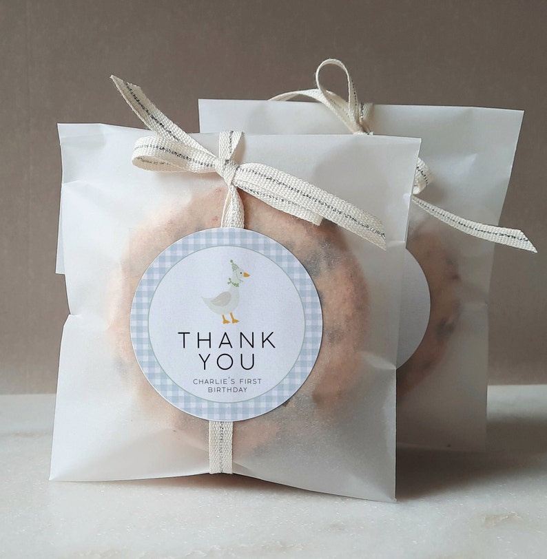 May include: White favor bags with a blue and white gingham circle sticker with a cartoon goose wearing a party hat. The sticker says "Thank You" and "Charlie's First Birthday".