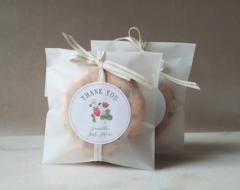 May include: Two white favor bags tied with a white and gray ribbon. Each bag contains a round cookie with a pink and white sticker that says "Thank You" with a strawberry illustration and the text "Smoother Baby Skin".