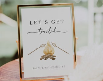 May include: A gold-framed sign with the text "Let's Get toasted" and a watercolor illustration of a campfire with marshmallows on sticks. The sign also includes the text "Sarah's Bachelorette".