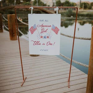May include: A white sign with red, white and blue American flag graphics and the text "Our All American Girl Ellie is One! 7/5/20XX". The sign is attached to a copper pipe stand.