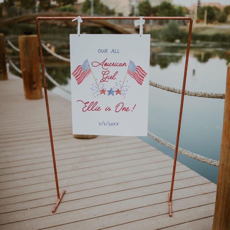 May include: A white sign with red, white and blue American flag graphics and the text "Our All American Girl Ellie is One! 7/5/20XX". The sign is attached to a copper pipe stand.