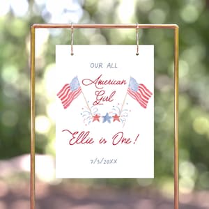 May include: A white sign with red, white and blue American flags and fireworks. The text reads "Our All American Girl Ellie is One! 7/3/20XX".