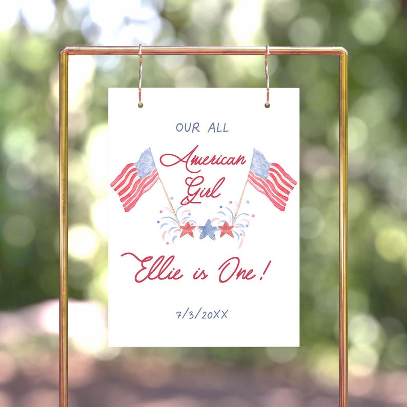 May include: A white sign with red, white and blue American flags and fireworks. The text reads "Our All American Girl Ellie is One! 7/3/20XX".