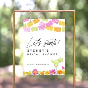 Op de afbeelding: Een wit bord met kleurrijke papieren vlaggetjes bovenaan. De tekst luidt "Let's fiesta! Sydney's Bridal Shower 02.11.23 | Wimberley, TX". Een margarita-glas met een limoenwig is onder de tekst afgebeeld.