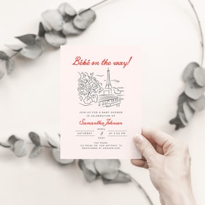 Op de afbeelding: Een roze babyborrel uitnodiging met een zwart-witte illustratie van de Eiffeltoren en bloemen. De tekst "B&eacute;b&eacute; on the way!" is rood. De uitnodiging is voor Samantha Johnson op zaterdag 3 april 2023 om 14:00 uur in The Pearl, 123 Pearl Dr. San Antonio, TX. De uitnodiging zegt "Registered at Amazon.com".