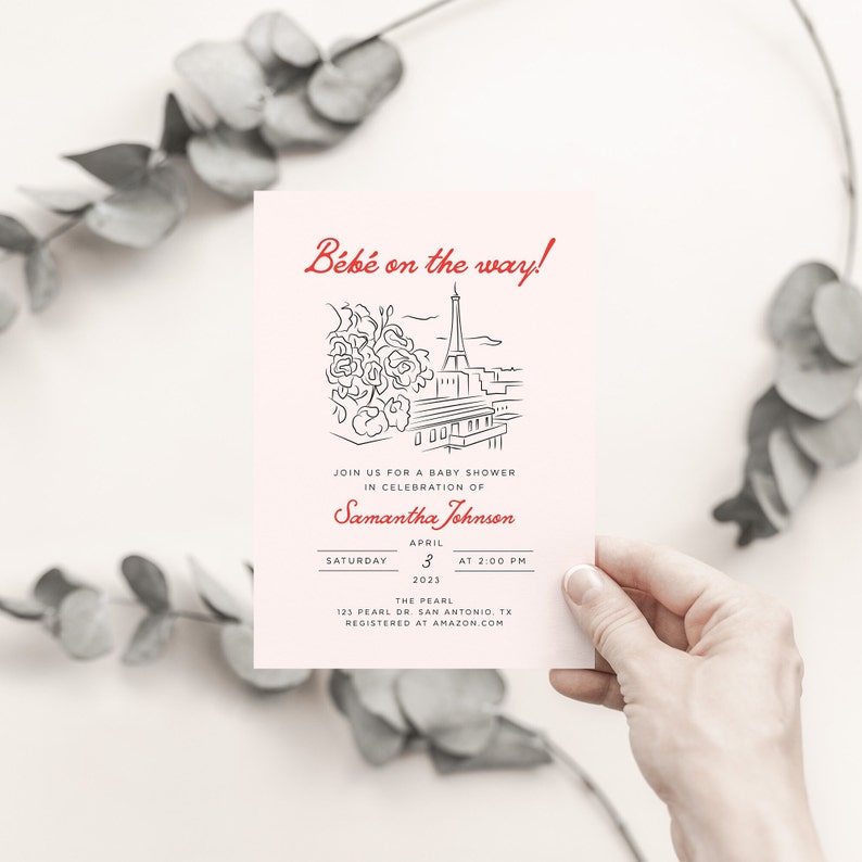 Op de afbeelding: Een roze babyborrel uitnodiging met een zwart-witte illustratie van de Eiffeltoren en bloemen. De tekst "B&eacute;b&eacute; on the way!" is rood. De uitnodiging is voor Samantha Johnson op zaterdag 3 april 2023 om 14:00 uur in The Pearl, 123 Pearl Dr. San Antonio, TX. De uitnodiging zegt "Registered at Amazon.com".