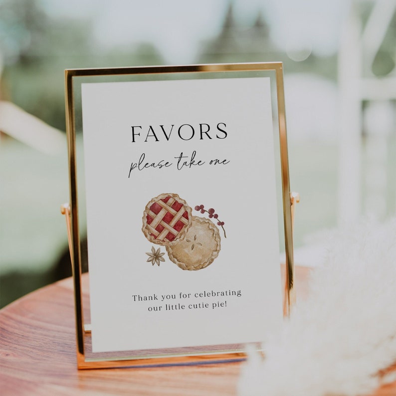 May include: A gold-framed sign with the text "Favors please take one" and a watercolor illustration of two pies. The text below the illustration reads "Thank you for celebrating our little cutie pie!"