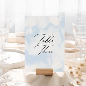 May include: A light blue table number card with a white watercolor background and the text "Table Three" in black calligraphy.