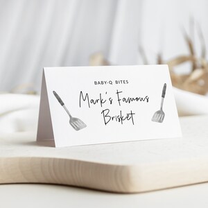May include: White card with black text that reads "BABY-Q BITES Mark's Famous Brisket" with two black and silver spatula illustrations on either side of the text.