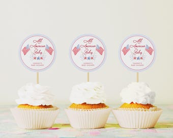 May include: Three cupcakes with white frosting and red, white, and blue cupcake toppers that say "All American Baby" with stars and fireworks. The toppers are for a baby shower.
