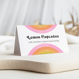 May include: A white card with a pink, orange, and yellow rainbow design. The text on the card reads "Lemon Cupcakes with strawberry buttercream frosting".