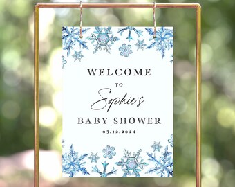 Puede incluir: Un letrero blanco con copos de nieve azules de acuarela y el texto "WELCOME to Sophie's BABY SHOWER 03.12.2024". El letrero está suspendido de un marco de metal dorado.