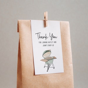 May include: A white thank you card with black text that reads "Thank You for joining Hayley and John's Baby-Q". A green and brown illustration of a grill is below the text. The card is attached to a brown paper bag with a wooden clothespin.