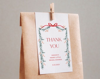 May include: A white gift tag with a red bow and the words "THANK YOU" in red. The tag is decorated with green and peach-colored floral elements and is clipped to a brown paper bag with a wooden clip. The text "Maggie's Merry Little Bridal Shower" and the date "8.22.2023" are also visible.