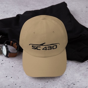 以下が含まれることがあります： 黒の刺繍で車のシルエットと「SC 430」の文字がデザインされた、タン色のベースボールキャップです。