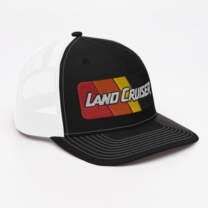 Puede incluir: Gorra de camionero negra y blanca con un parche a rayas rojo, naranja, amarillo y blanco que dice "LAND CRUISER" en letras blancas.