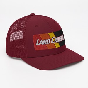 Puede incluir: Una gorra de camionero color burdeos con un parche a rayas negro y amarillo que dice "LAND CRUISER" en letras blancas.