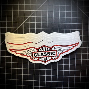 May include: A stack of stickers featuring a stylized wing design with red accents and the words "AIR CLASSIC COOLED" in a rectangular emblem. The stickers are white with a glossy finish, set against a black grid background.