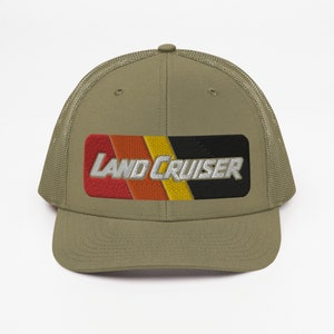 Puede incluir: Una gorra de camionero verde caqui con una parte trasera de malla negra. La parte delantera de la gorra presenta un parche a rayas rojo, naranja, amarillo y negro con el texto "LAND CRUISER" en blanco.