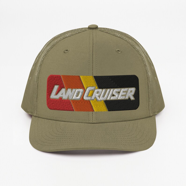 Puede incluir: Una gorra de camionero verde caqui con una parte trasera de malla negra. La parte delantera de la gorra presenta un parche a rayas rojo, naranja, amarillo y negro con el texto "LAND CRUISER" en blanco.
