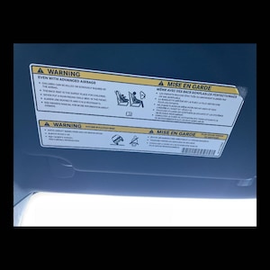 May include: A car sun visor displaying safety warnings in English and French. The warnings cover airbags, child safety, and rollover risks. The visor is a light grey colour.