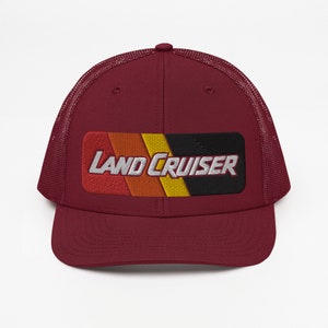 Puede incluir: Una gorra de camionero granate con una malla negra en la parte posterior. La parte delantera de la gorra presenta un parche a rayas rojo, naranja, amarillo y negro con el texto "LAND CRUISER" en blanco.