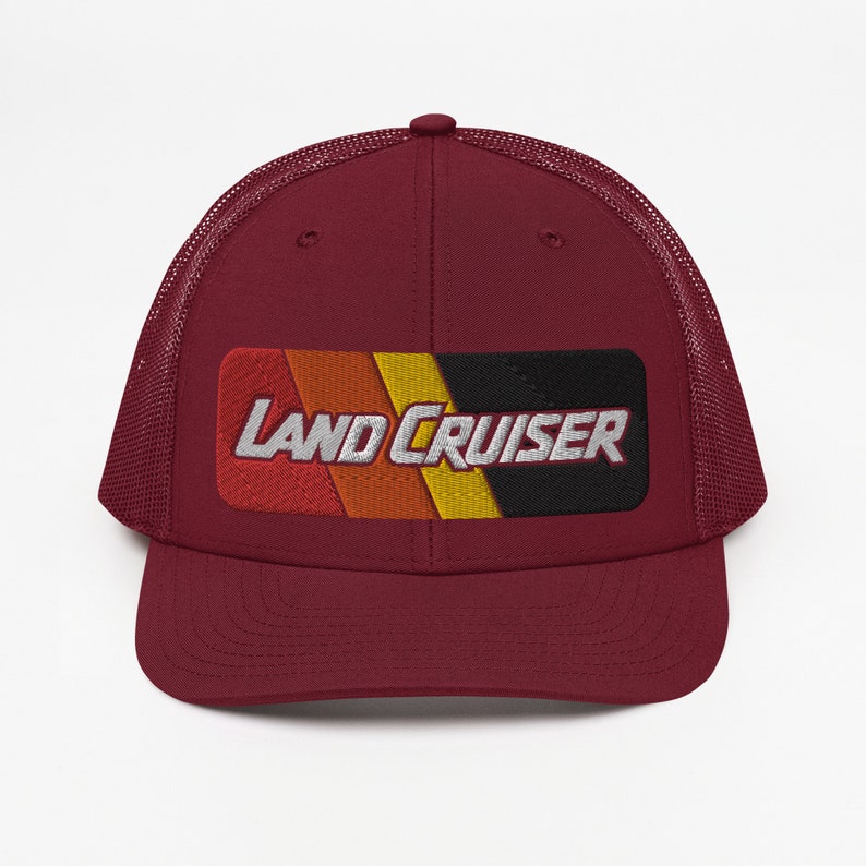 Puede incluir: Una gorra de camionero granate con una malla negra en la parte posterior. La parte delantera de la gorra presenta un parche a rayas rojo, naranja, amarillo y negro con el texto "LAND CRUISER" en blanco.