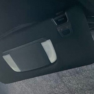 Puede incluir: Un parasol de coche negro con forma rectangular y un espejo de cortesía incorporado. El parasol tiene un panel rectangular de color claro que se puede bajar. El interior del coche es visible.