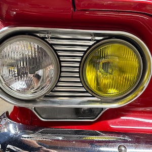 Könnte beinhalten: Nahaufnahme der Scheinwerferanordnung eines Oldtimers. Das Bild zeigt ein rotes Auto mit einer verchromten Scheinwerfereinfassung. Die Scheinwerfer sind rund, mit einer klaren und einer gelben Linse. Die Karosserie des Autos ist rot.