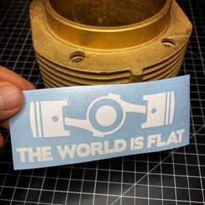 Op de afbeelding: Een witte sticker met een afbeelding van motorzuigers en de tekst "THE WORLD IS FLAT" wordt voor een goudkleurige motorcilinder gehouden. De sticker staat op een blauwe achtergrond.