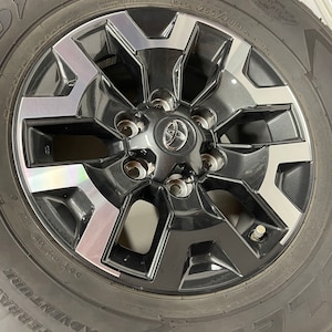 May include: A Goodyear Wrangler All-Terrain Adventure tire mounted on a black and silver alloy wheel. The wheel has a Toyota center cap and is marked with tire size 265/70R18.